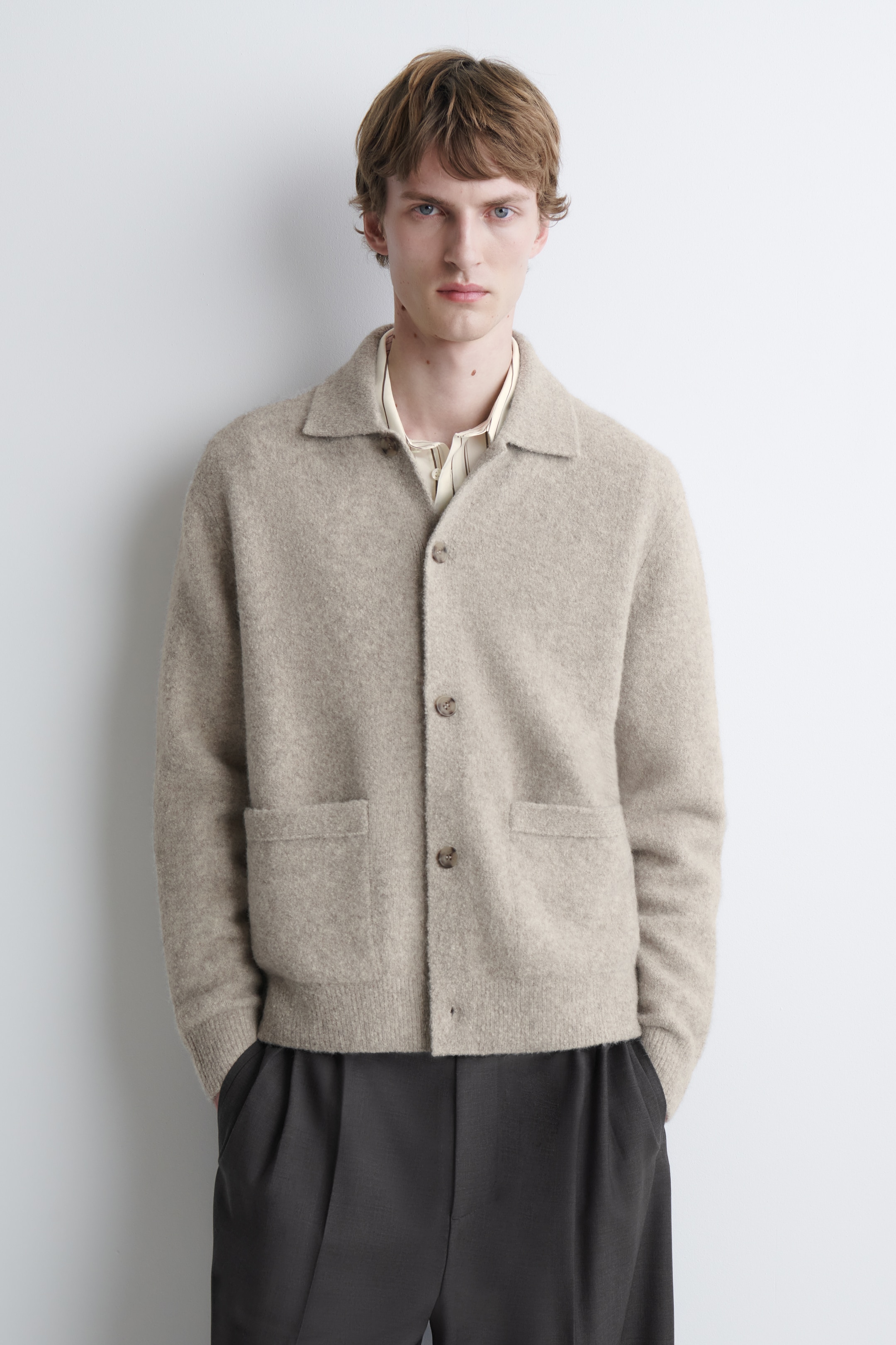 Person wearing a beige wool cardigan over a cream shirt and dark trousers, standing against a white background.