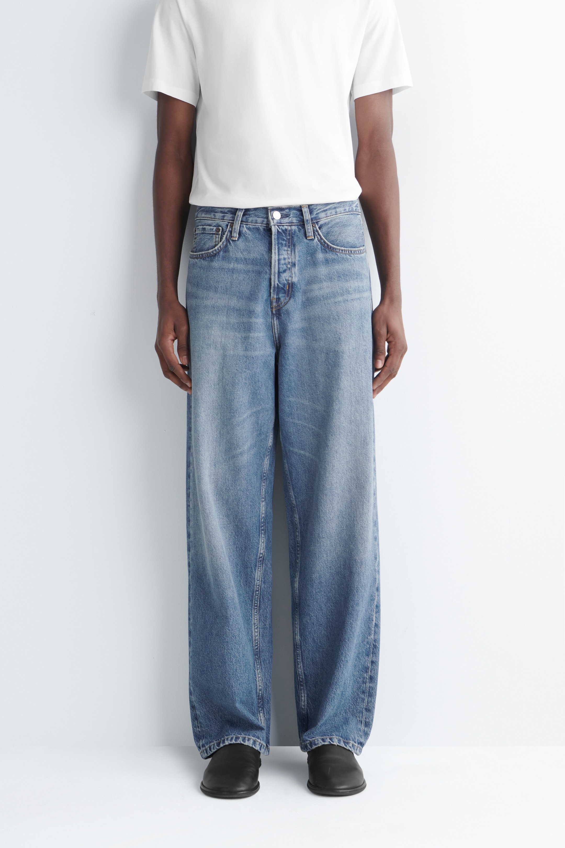 Person wearing a plain white t-shirt, loose blue jeans, and black shoes, standing in front of a white background.