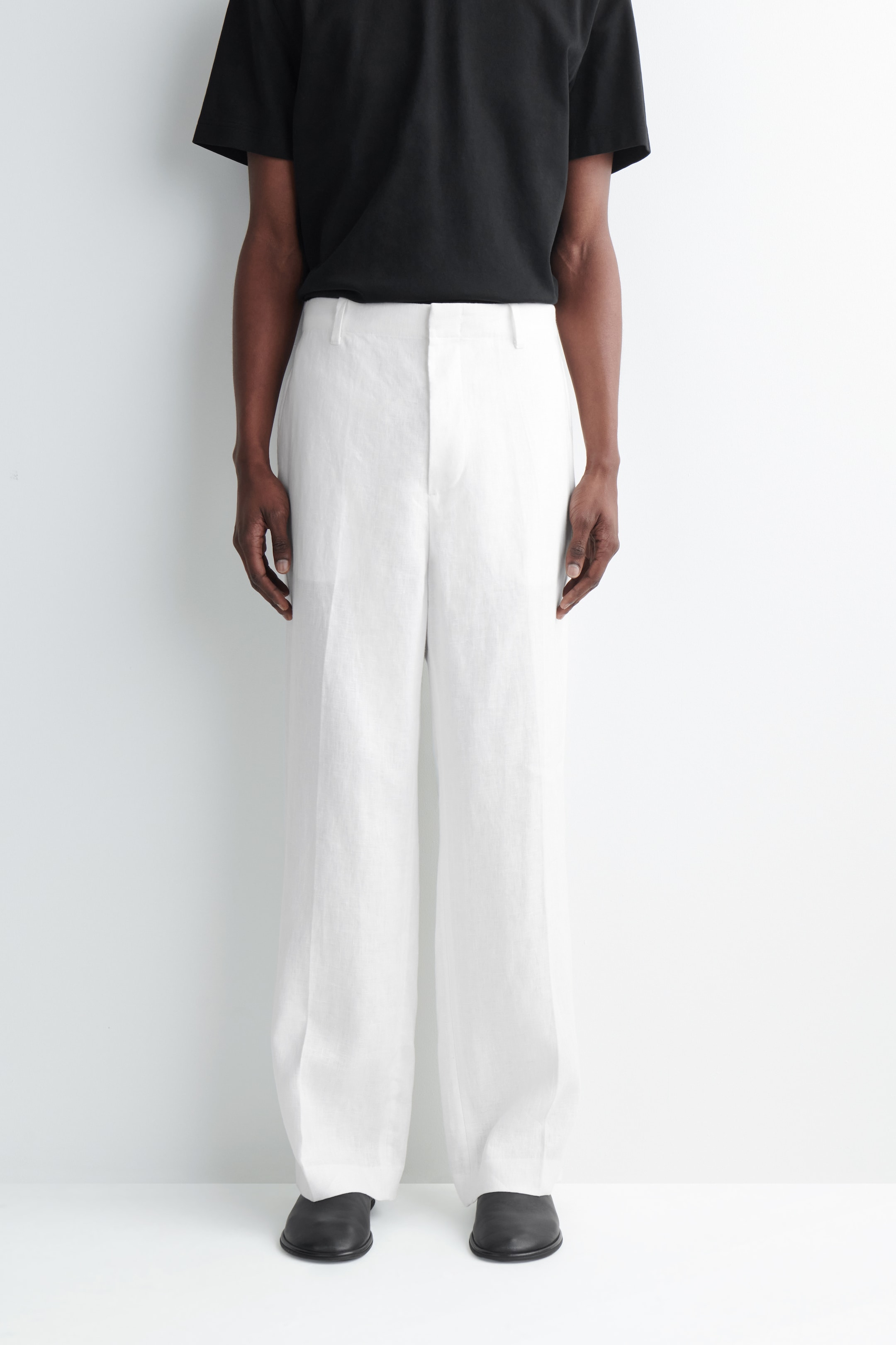 Person wearing a black T-shirt, white wide-leg trousers, and black shoes, standing against a plain white background.