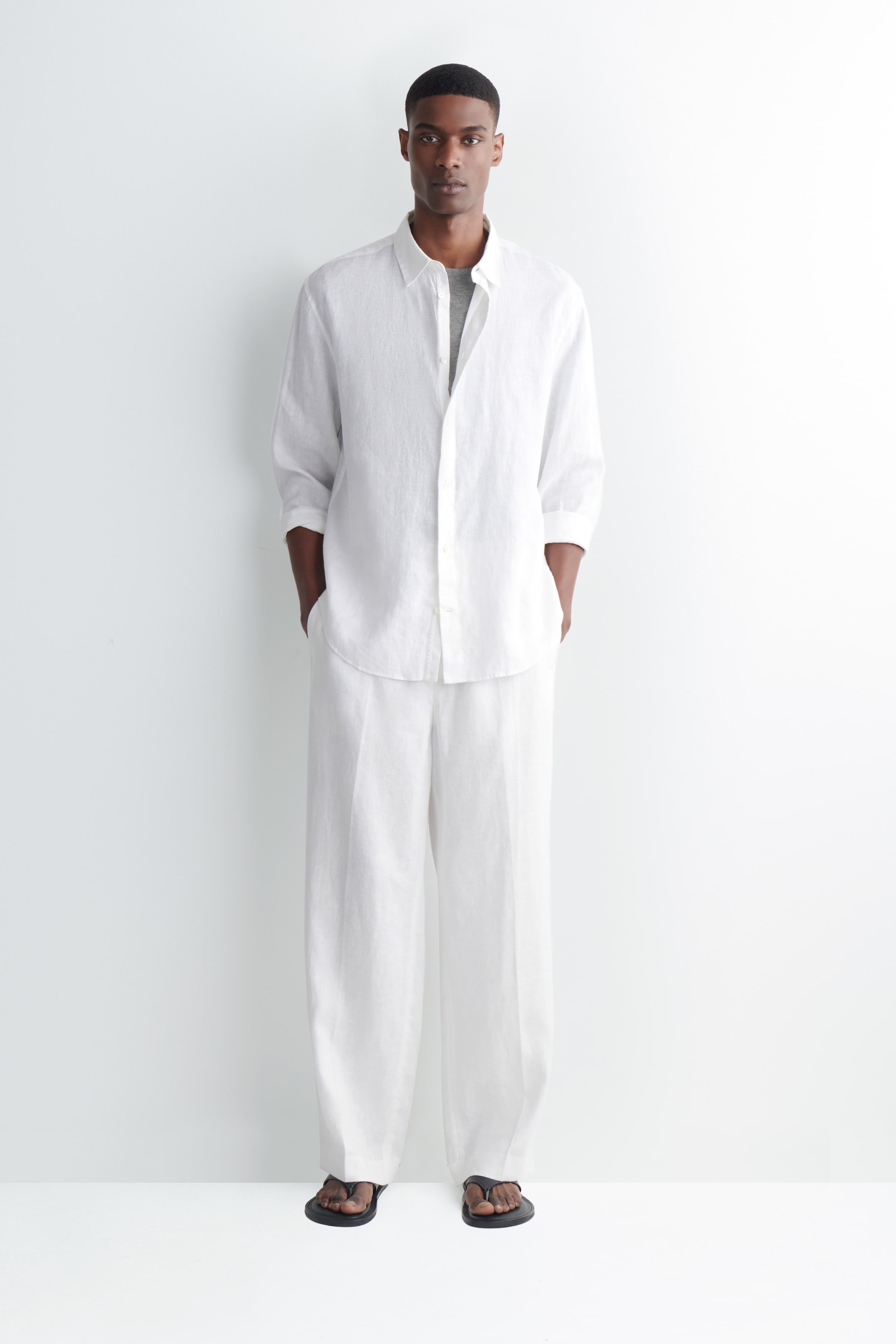 Man in a white linen shirt and pants stands against a white background, hands in pockets, wearing black sandals.