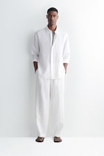 Placeholder for: Man in a white linen shirt and pants stands against a white background, hands in pockets, wearing black sandals.