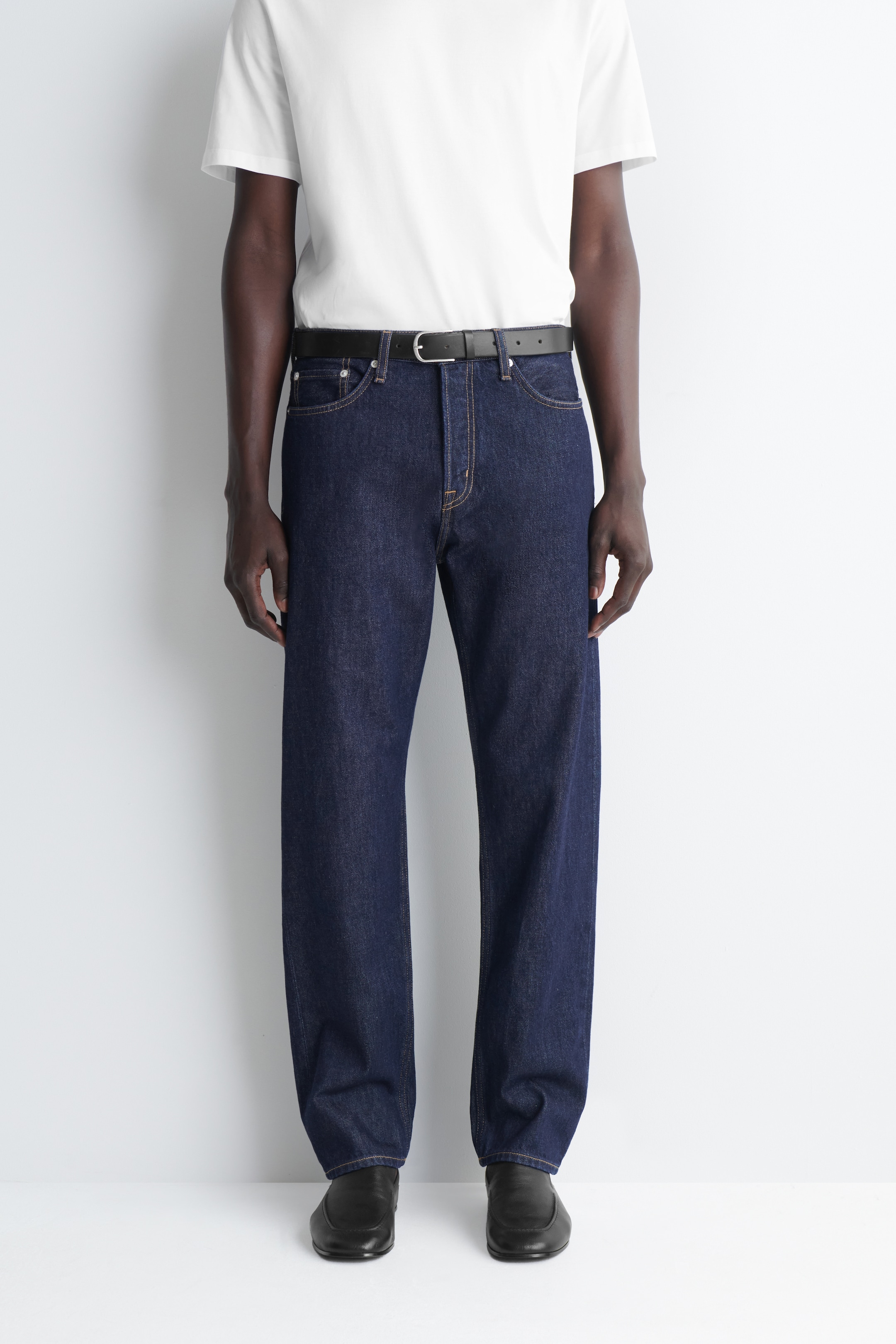 Man wearing dark blue jeans, a white T-shirt, black belt, and black shoes, standing against a white background.
