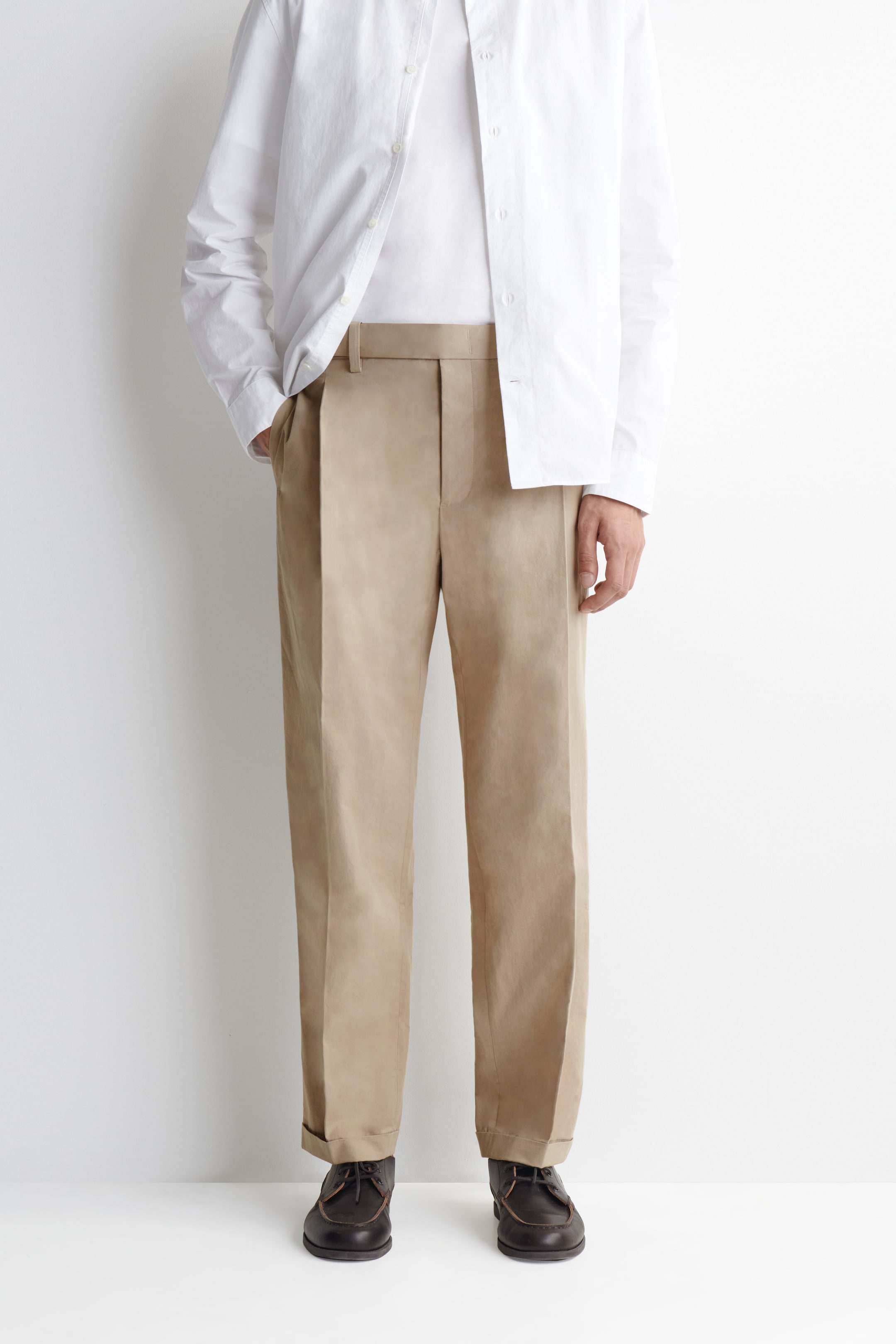 A person wearing a white shirt, beige wide-leg trousers, and black shoes, standing against a plain white background.