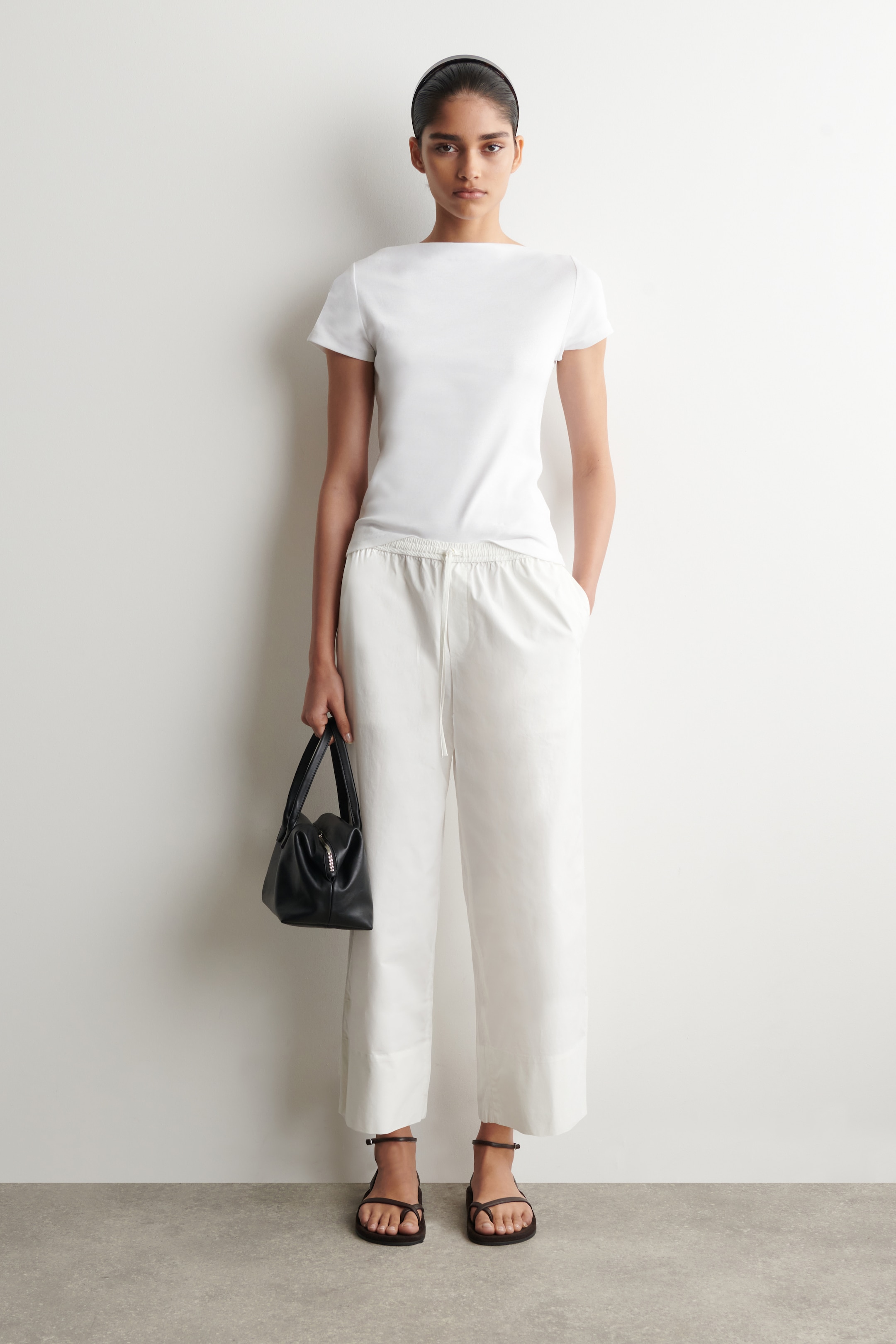 Person in a white T-shirt and pants, holding a black handbag, stands against a plain background wearing black sandals.