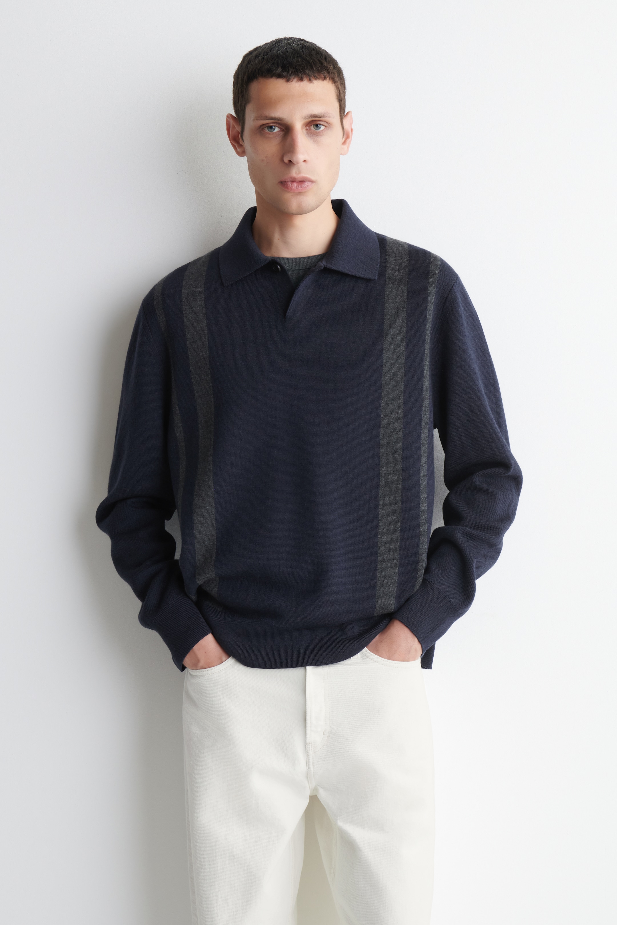 Person wearing a navy sweater with vertical gray stripes and white pants, standing with hands in pockets against a plain white background.