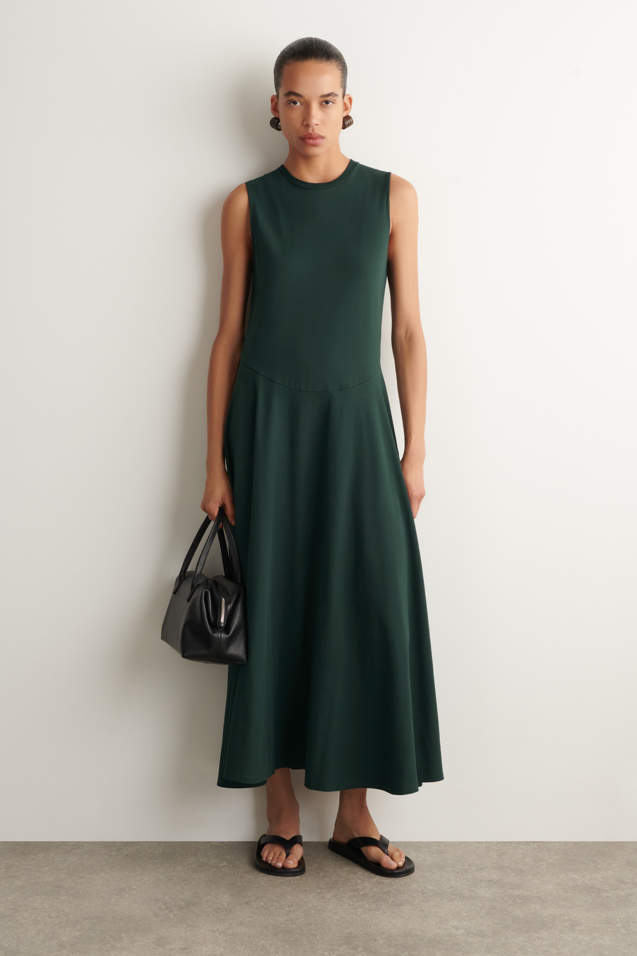 A person in a sleeveless dark green dress holds a black handbag, wearing flip-flops and large earrings, standing against a white wall.