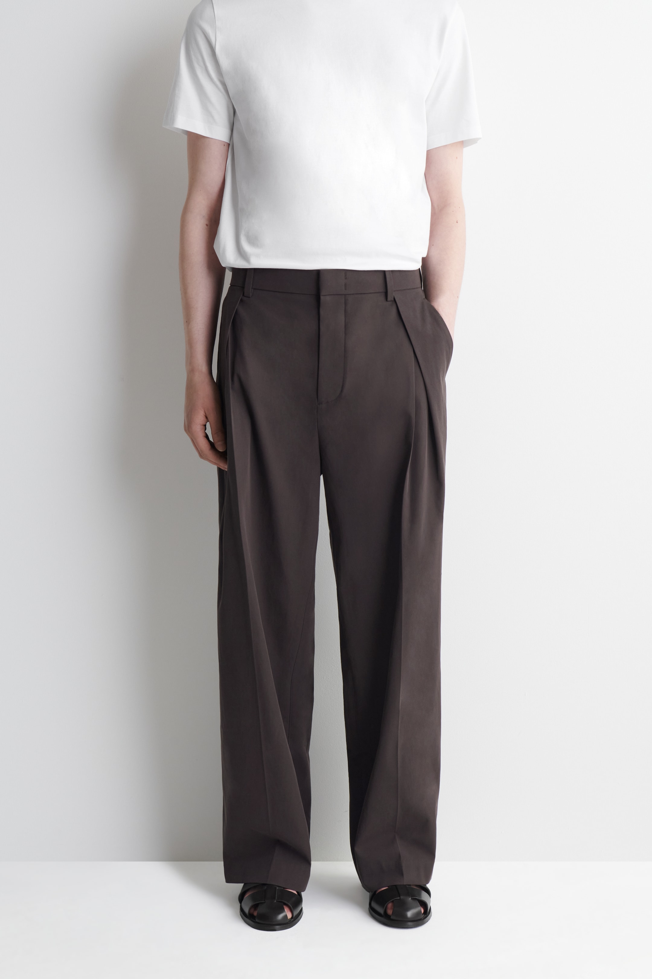 Person wearing a white T-shirt, brown pleated trousers, and black sandals, standing against a white background.