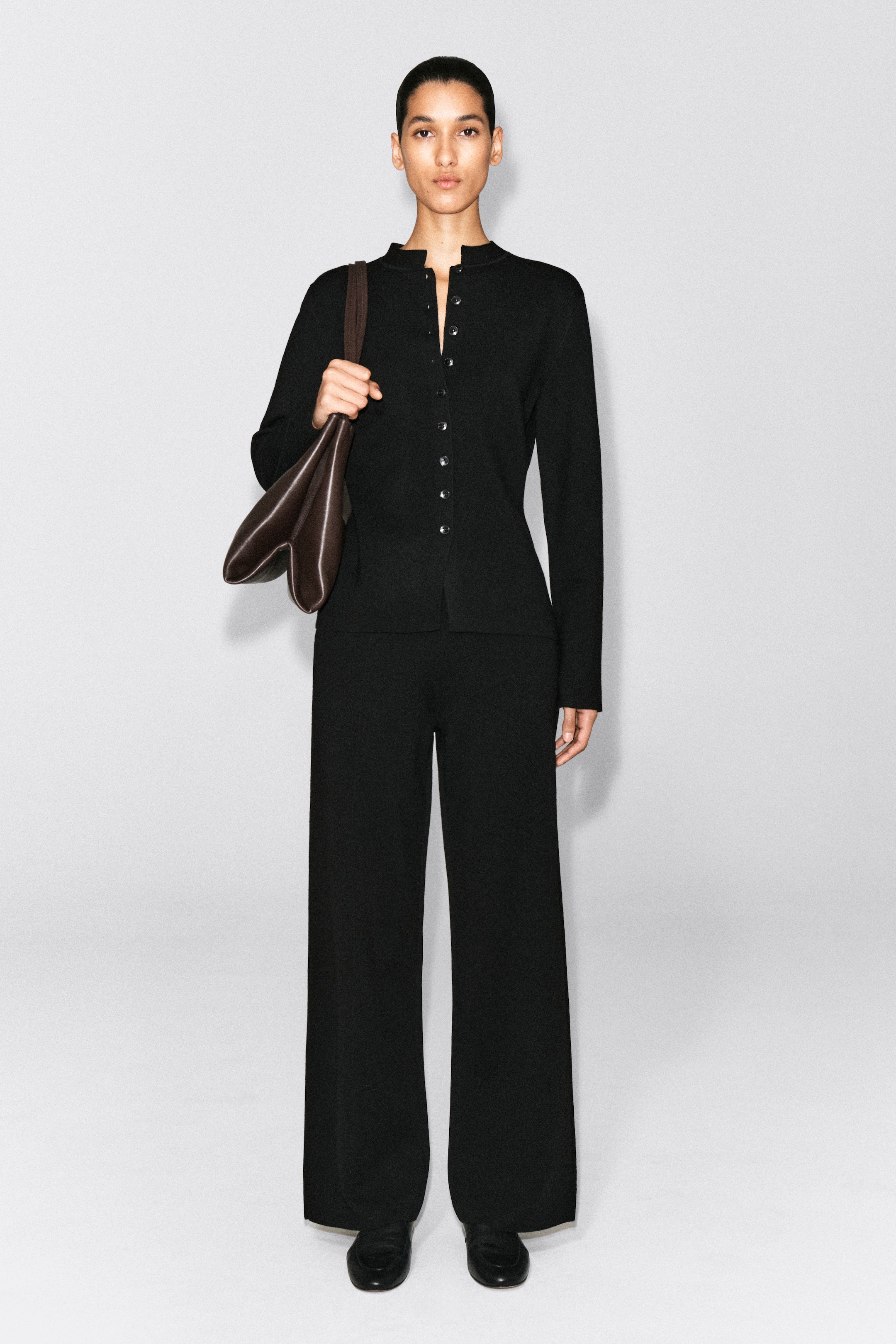 A person in a black button-up outfit holds a brown bag, standing against a plain white background.