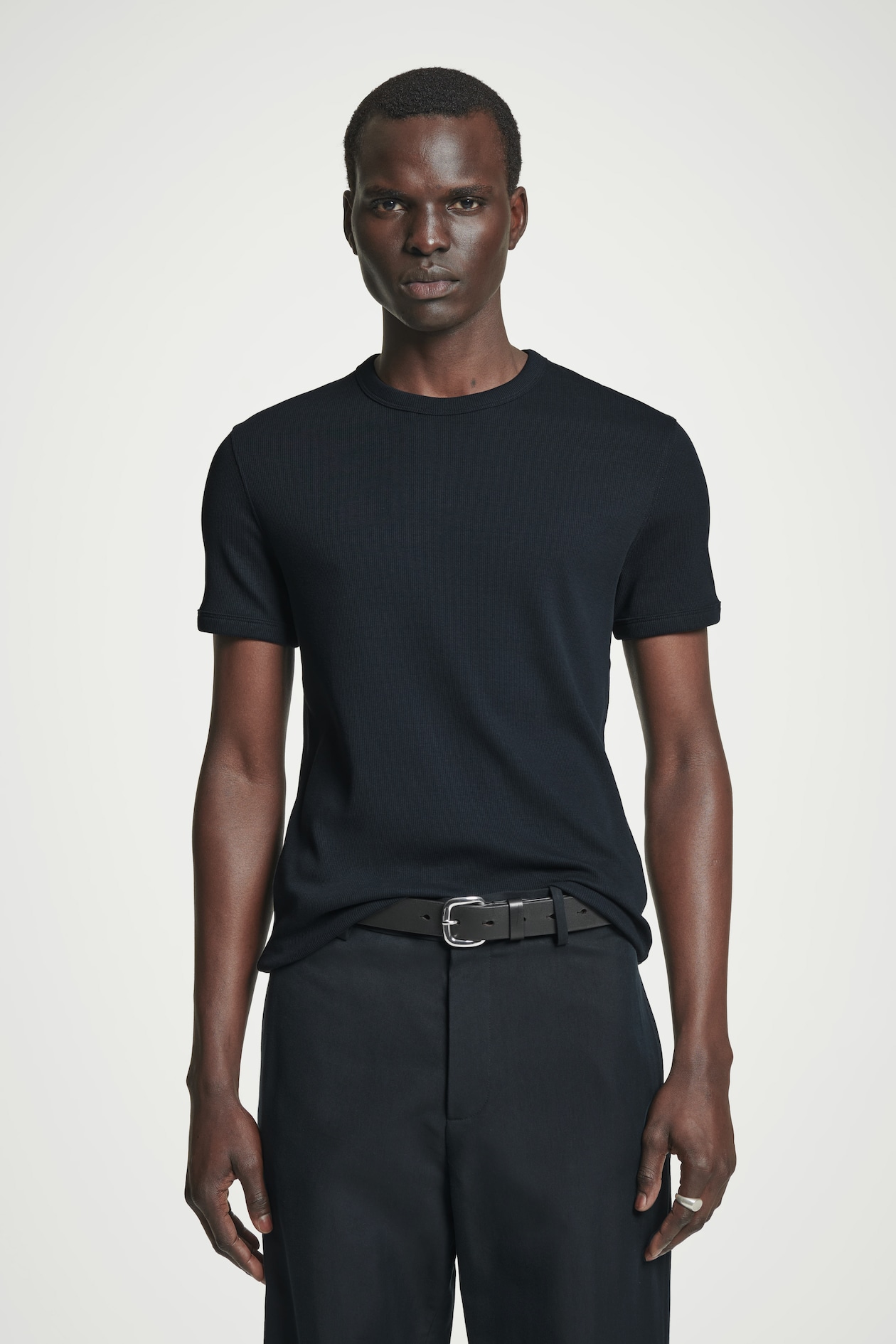 Person wearing a fitted black t-shirt and black pants with a belt, standing against a plain white background.