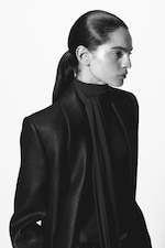 Placeholder for: A person in a sleek, dark outfit with a high collar and scarf, hair tied back, gazes to the side against a plain background.