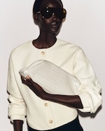 Placeholder for: Person wearing oversized sunglasses and a cream-colored jacket, holding a large white textured clutch bag against a plain background.