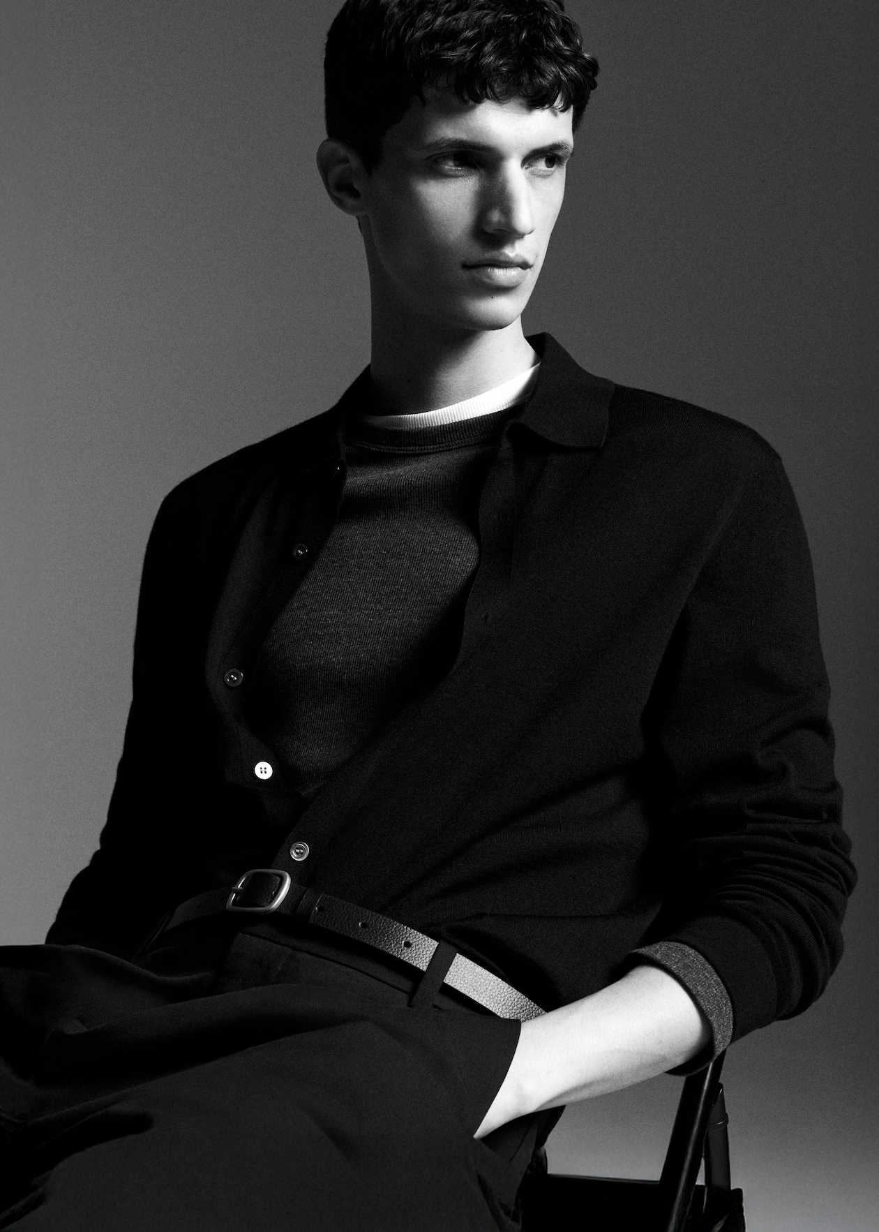 Black and white portrait of a person sitting, wearing a dark sweater and pants, looking to the side, with a neutral expression.