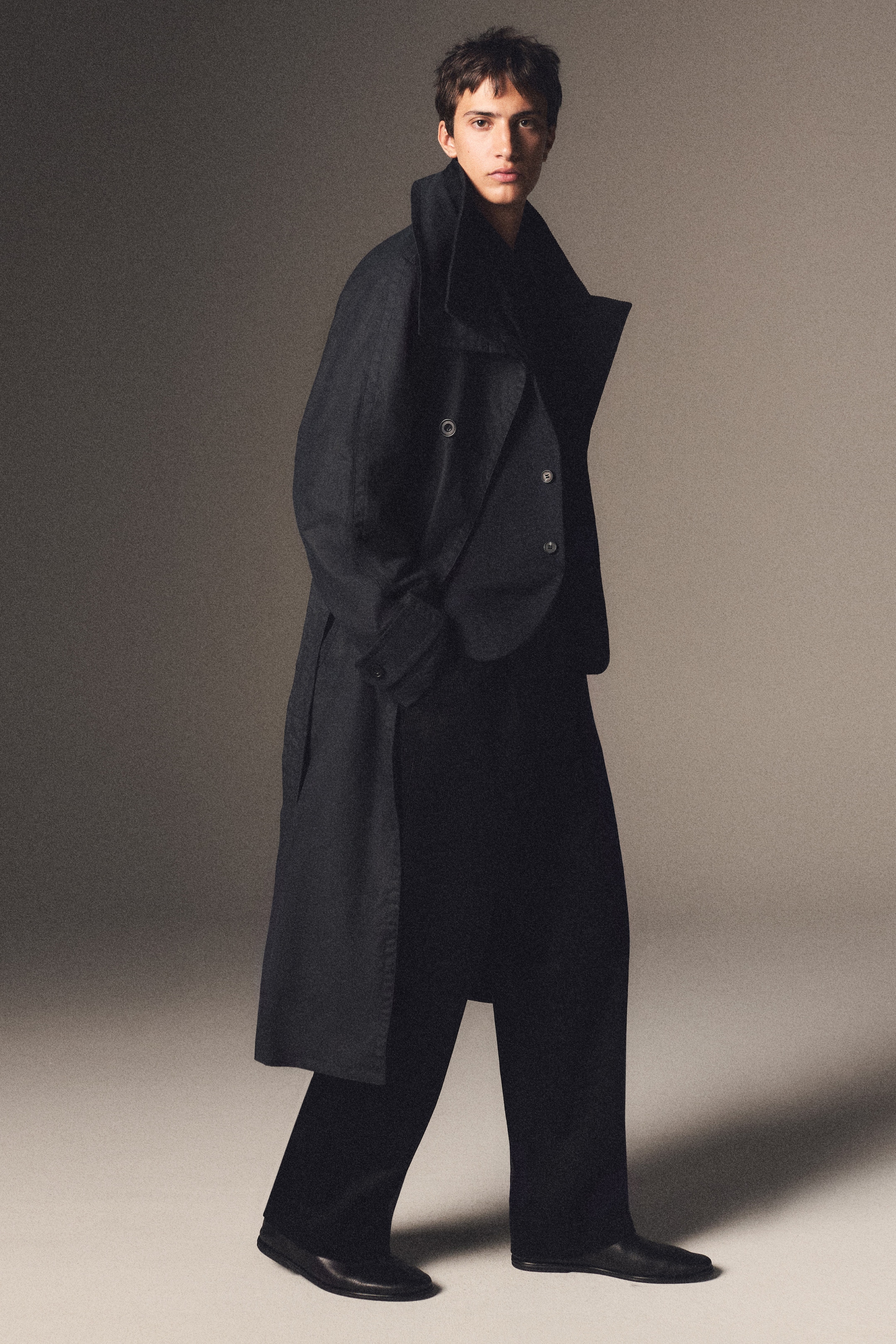 A person wearing a long, dark coat and black trousers, standing in a neutral-toned studio, looking forward.