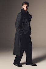 Placeholder for: A person wearing a long, dark coat and black trousers, standing in a neutral-toned studio, looking forward.