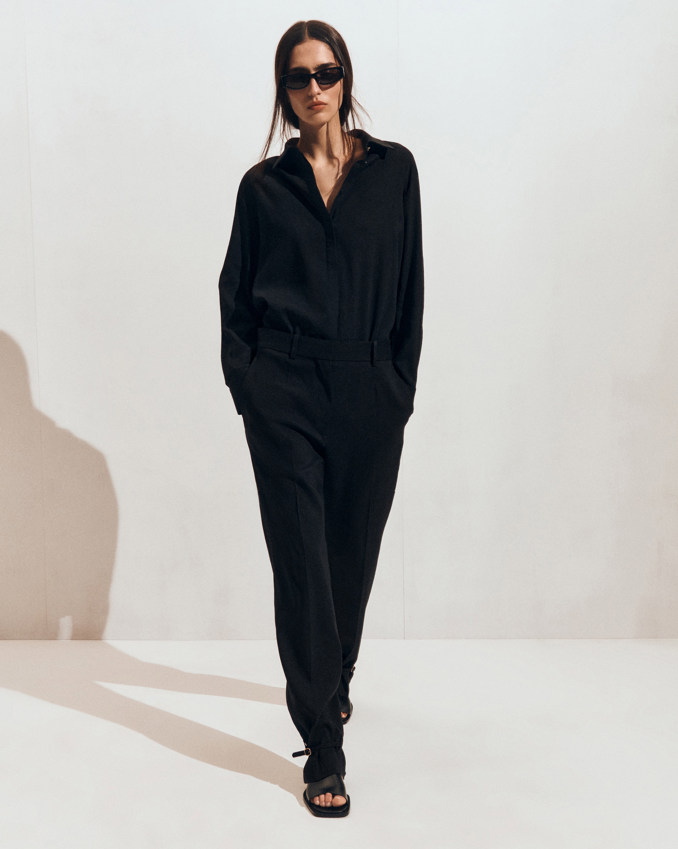 Woman wearing a black outfit, sunglasses, and sandals, walking confidently against a neutral background.