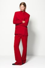 Placeholder for: Person in a bold red suit with a high collar poses against a white background, arms crossed, exuding confidence and style.