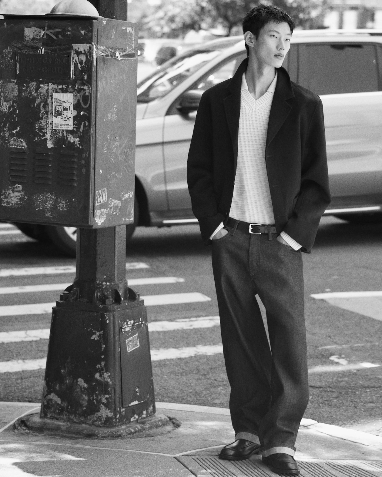 A person in a stylish outfit stands on a city sidewalk near a utility box, with a car passing by in the background.