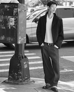 Placeholder for: A person in a stylish outfit stands on a city sidewalk near a utility box, with a car passing by in the background.