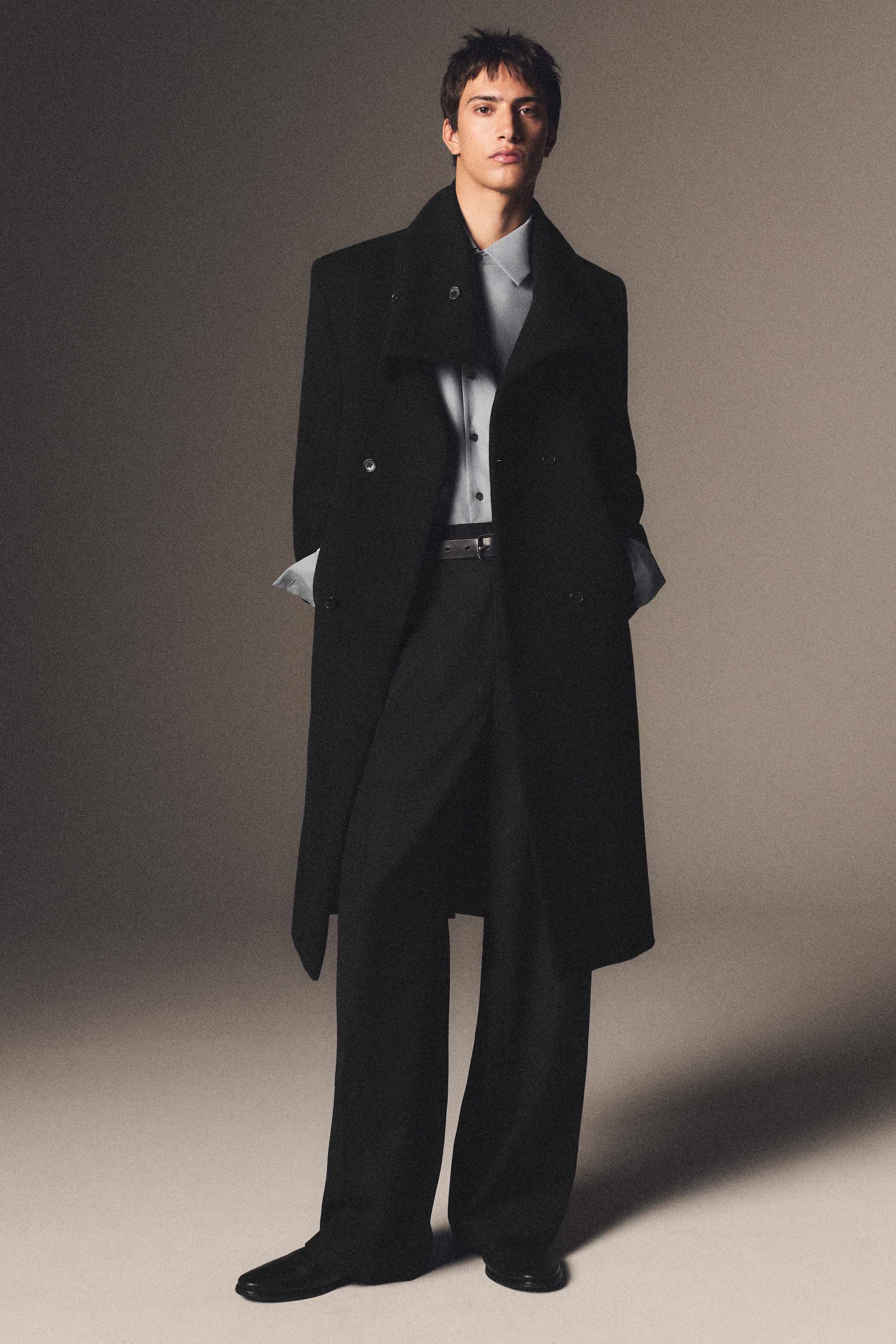 Person wearing an elegant black overcoat over a light blue shirt and black trousers, standing with hands in pockets against a neutral background.