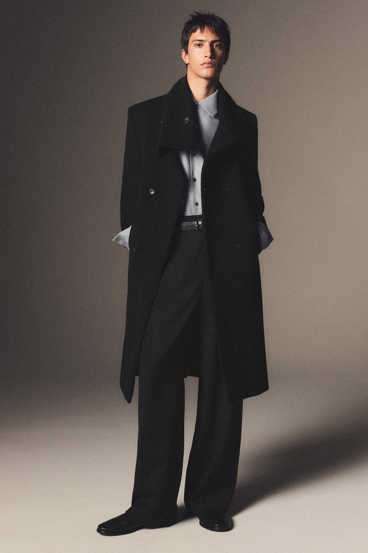 A person in a stylish black overcoat over a light-blue shirt and black trousers poses confidently against a neutral background.