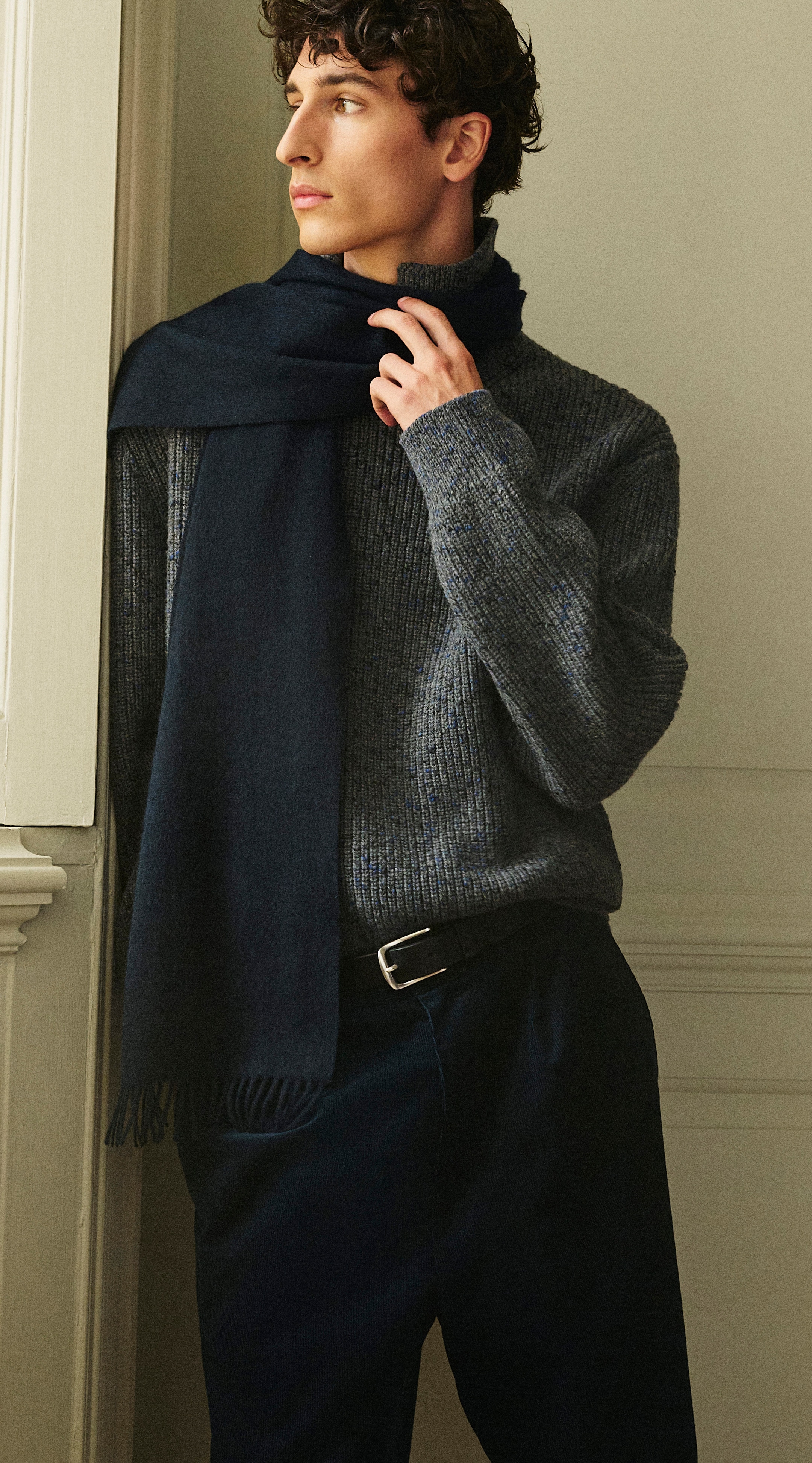Man in a gray sweater and dark blue scarf, gazing to the side by a window, with hands gently adjusting the scarf.
