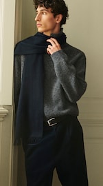Placeholder for: Man in a gray sweater and dark blue scarf, gazing to the side by a window, with hands gently adjusting the scarf.