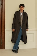 Placeholder for: Person wearing a dark long coat over a patterned vest and jeans, leaning against a wall in a room with wooden trim and white paneling.