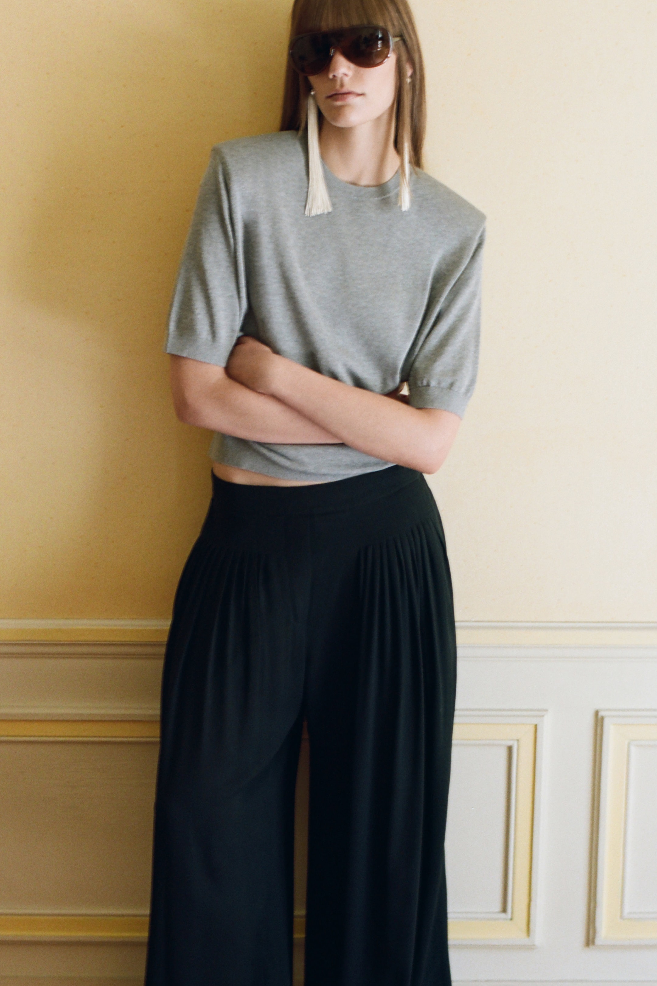 Person wearing sunglasses, gray top, and black wide-leg pants, standing against a light-colored wall with arms crossed.
