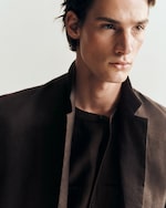 Placeholder for: Man in a stylish brown jacket and shirt, gazing intently to the side, with a neutral background.