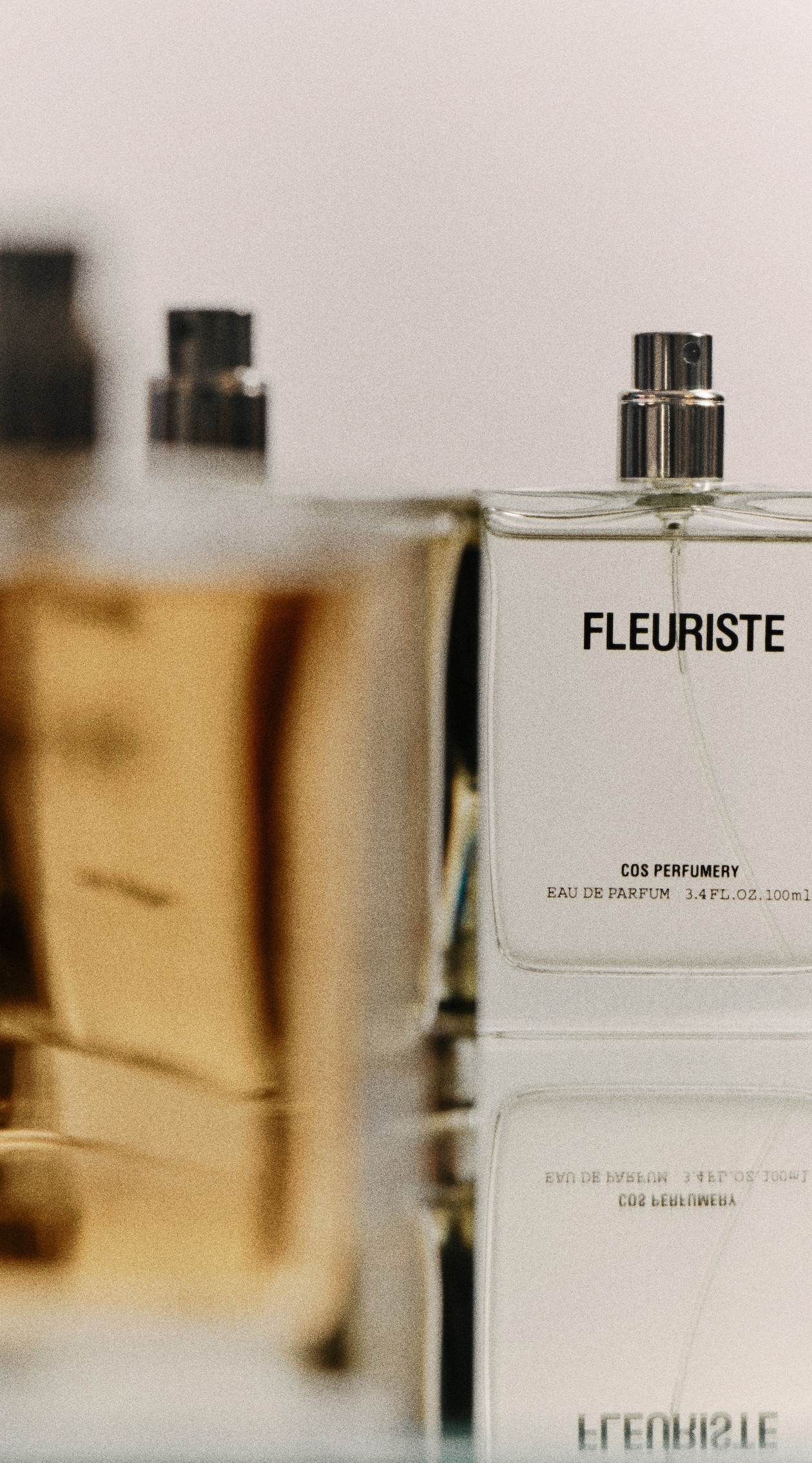 Close-up of a Fleuriste perfume bottle with text "COS Perfumery" and "Eau de Parfum" visible, reflected on a glossy surface.