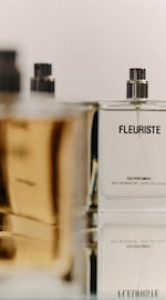 Placeholder for: Close-up of a Fleuriste perfume bottle with text "COS Perfumery" and "Eau de Parfum" visible, reflected on a glossy surface.