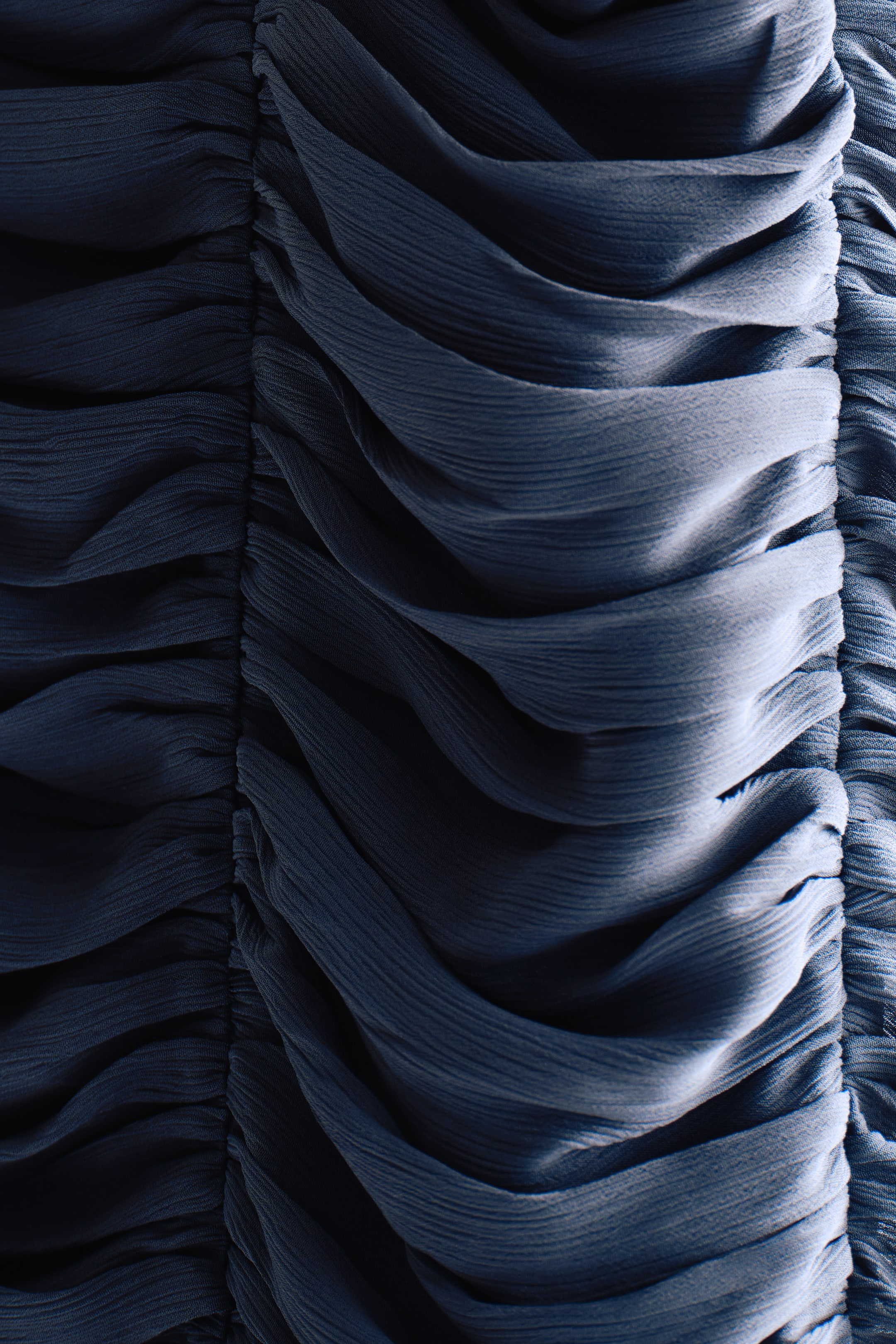 Close-up of dark blue, pleated fabric showcasing intricate textures and folds, with light creating subtle highlights.