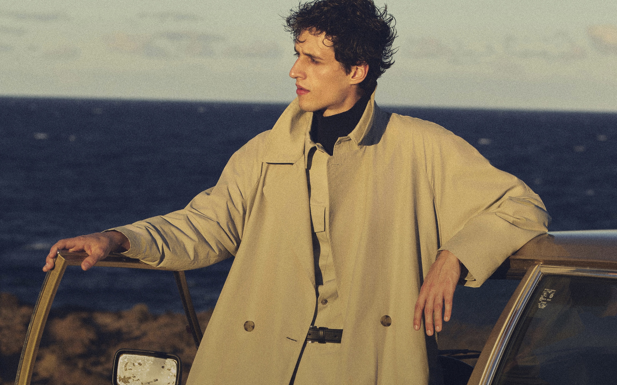 Person in a beige coat leans on a car door, gazing over the ocean at sunset.