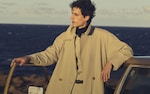 Placeholder for: Person in a beige coat leans on a car door, gazing over the ocean at sunset.