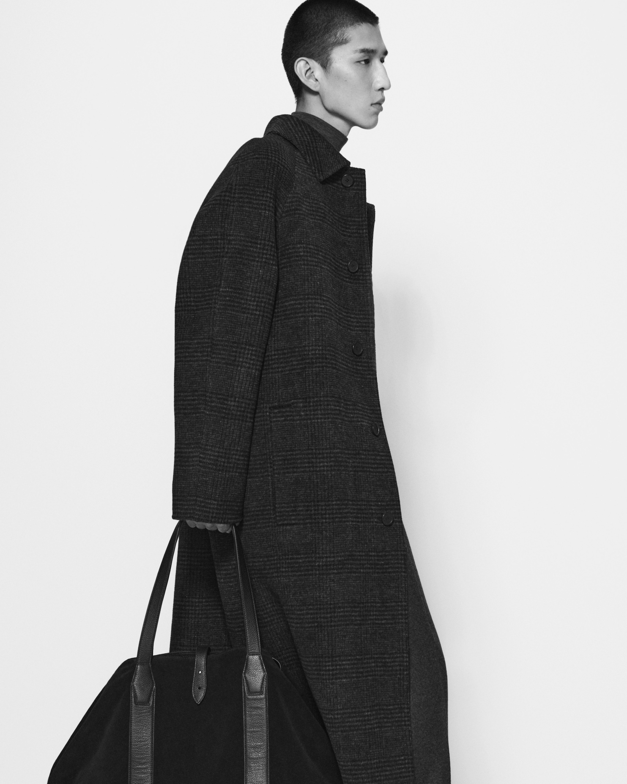 Person in a long, dark plaid coat carrying a large black bag, walking against a plain white background.