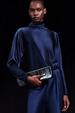 Placeholder for: Person wearing a dark blue satin outfit holds a textured black clutch against a black background.