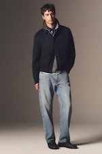 Placeholder for: A person wearing a dark jacket, light blue jeans, and black shoes, posing against a neutral background.