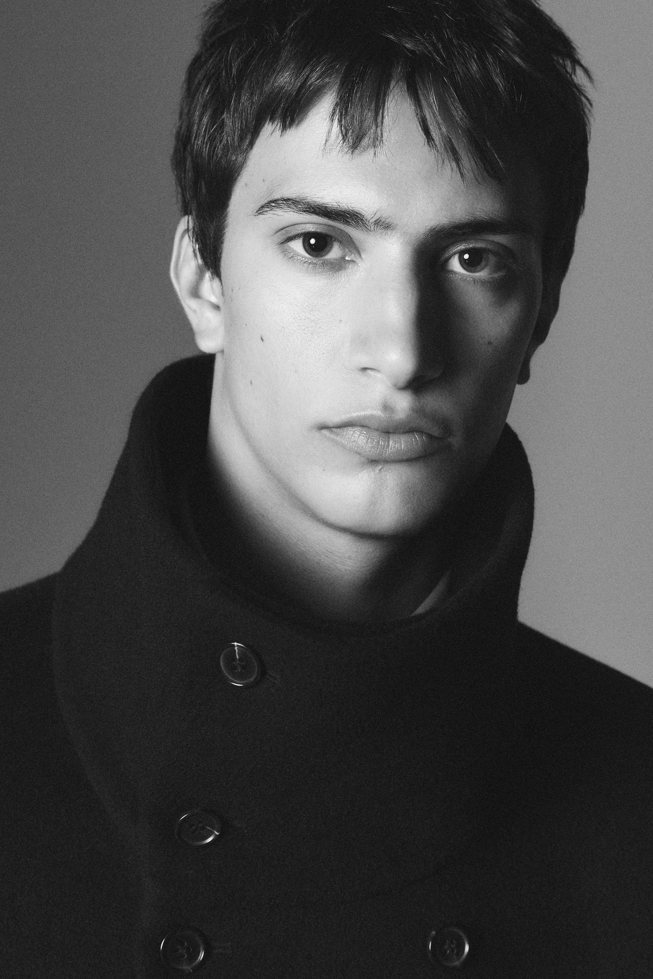 Black and white portrait of a person with short dark hair, wearing a high-collared coat, gazing directly at the camera.