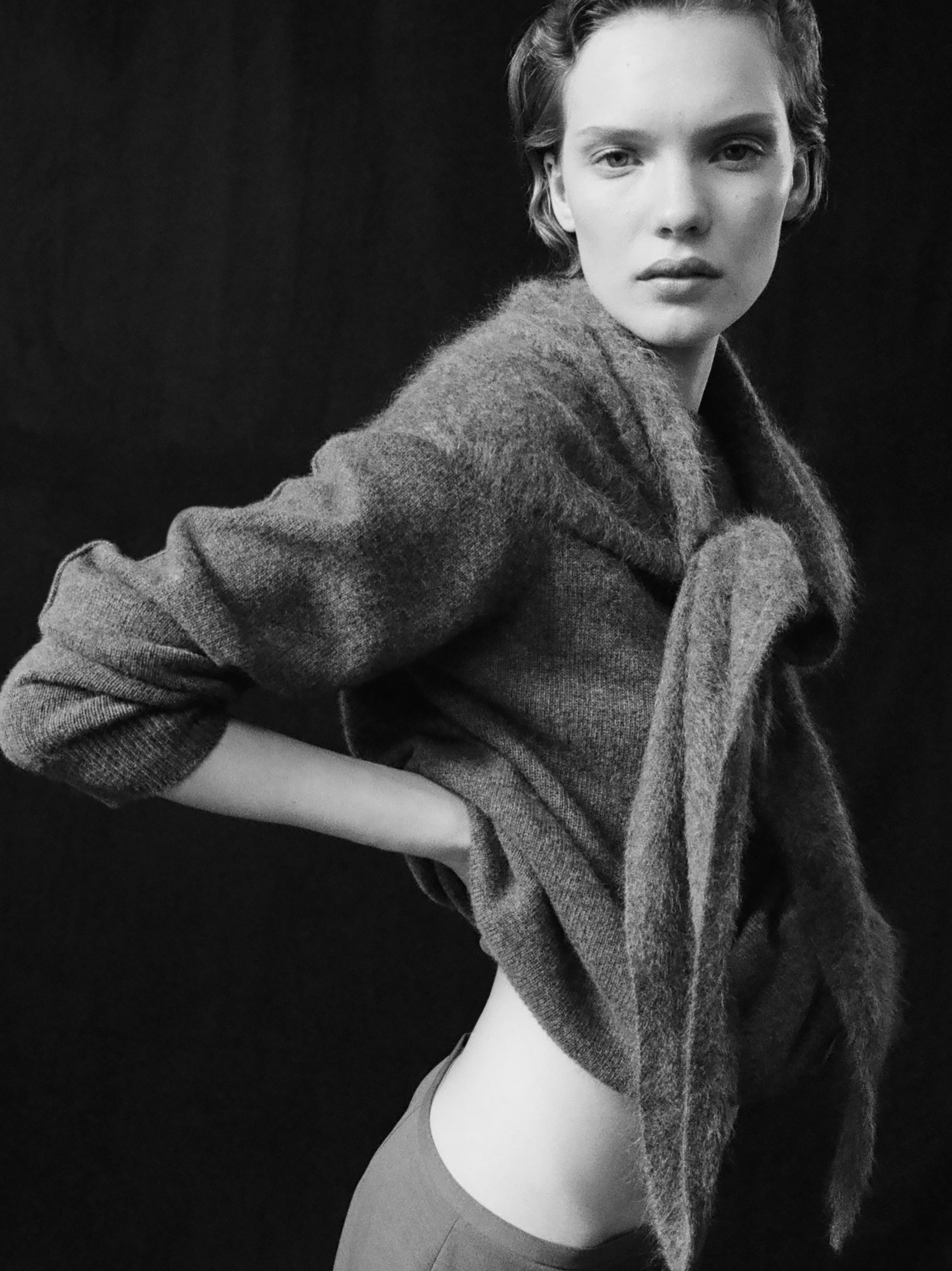 Black and white image of a person posing confidently, wearing a soft textured sweater, with a dark background.