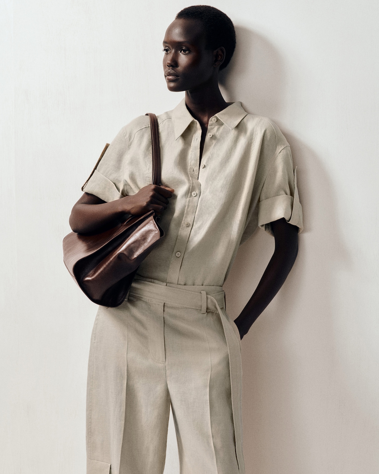 Person wearing a light beige short-sleeved button-up shirt and matching pants, holding a brown leather bag, standing against a plain backdrop.