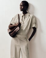 Placeholder for: Person wearing a light beige short-sleeved button-up shirt and matching pants, holding a brown leather bag, standing against a plain backdrop.
