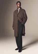 Placeholder for: A person models a stylish brown coat over a layered outfit, featuring a collared shirt, sweater, and black trousers, standing confidently.
