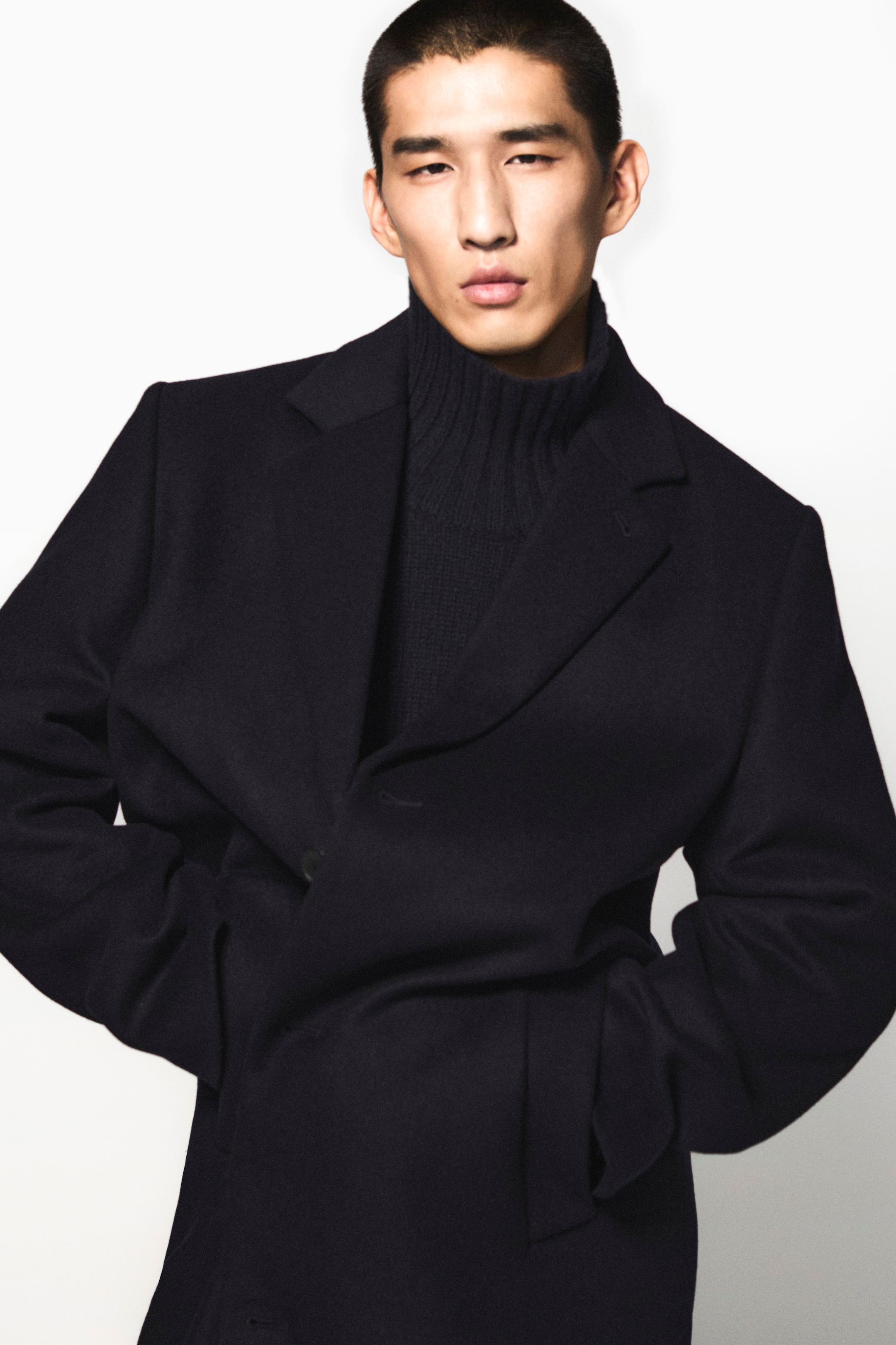 Person wearing a dark coat and turtleneck, standing against a plain white background.