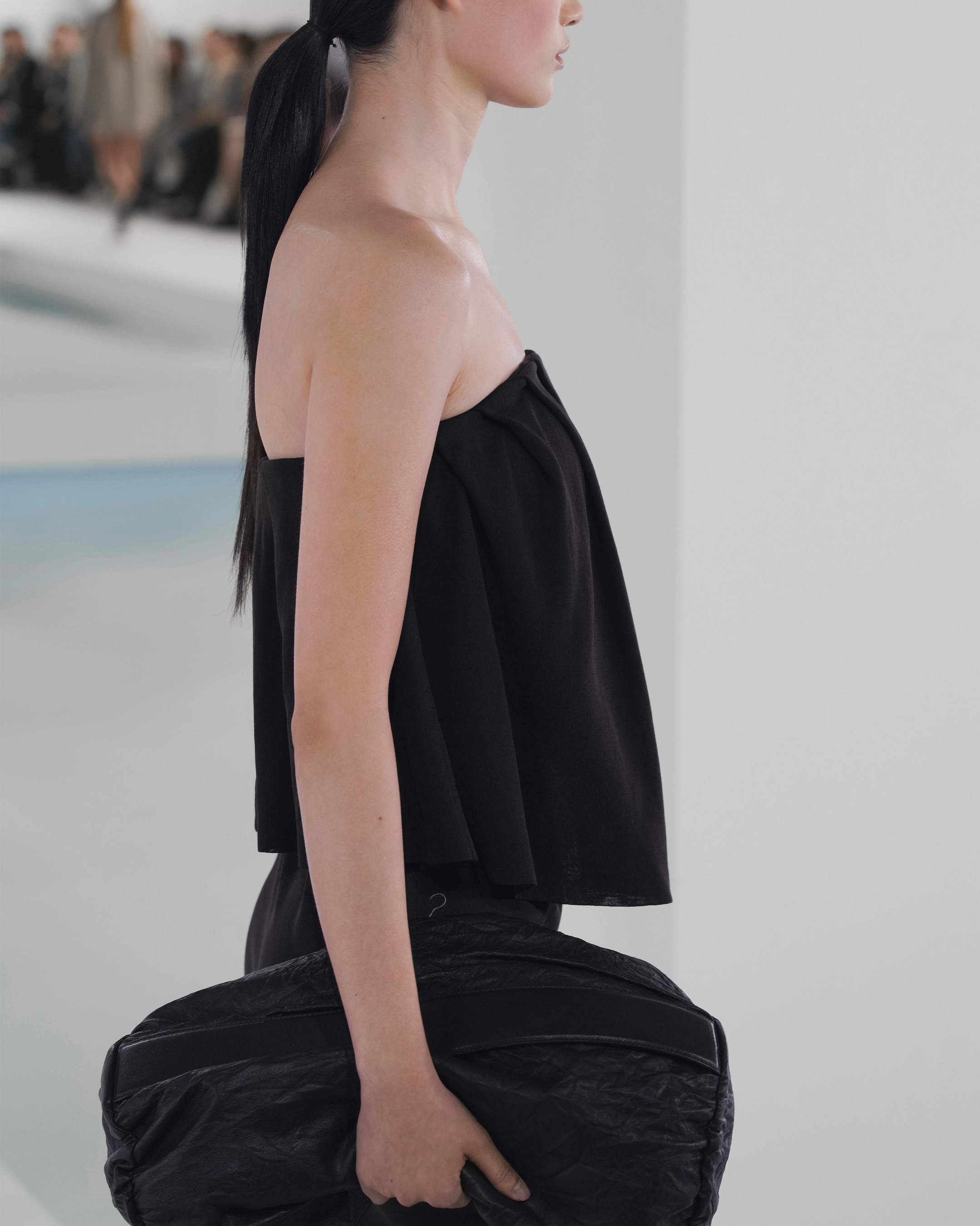 A model in a black strapless top and ponytail carries a large black bag on a runway.