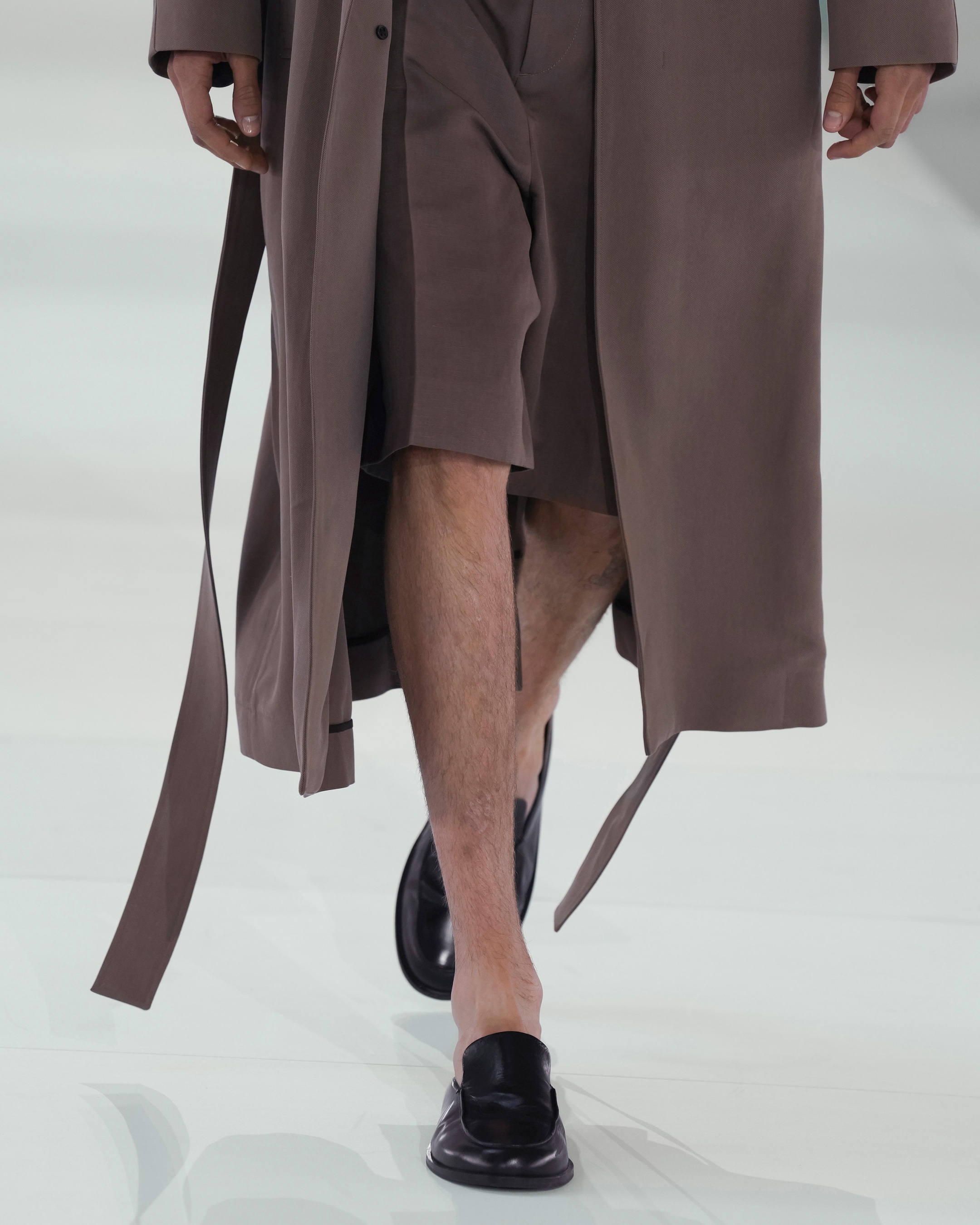 Person walking on a runway wearing a long brown coat, matching shorts, and black loafers.