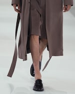 Placeholder for: Person walking on a runway wearing a long brown coat, matching shorts, and black loafers.