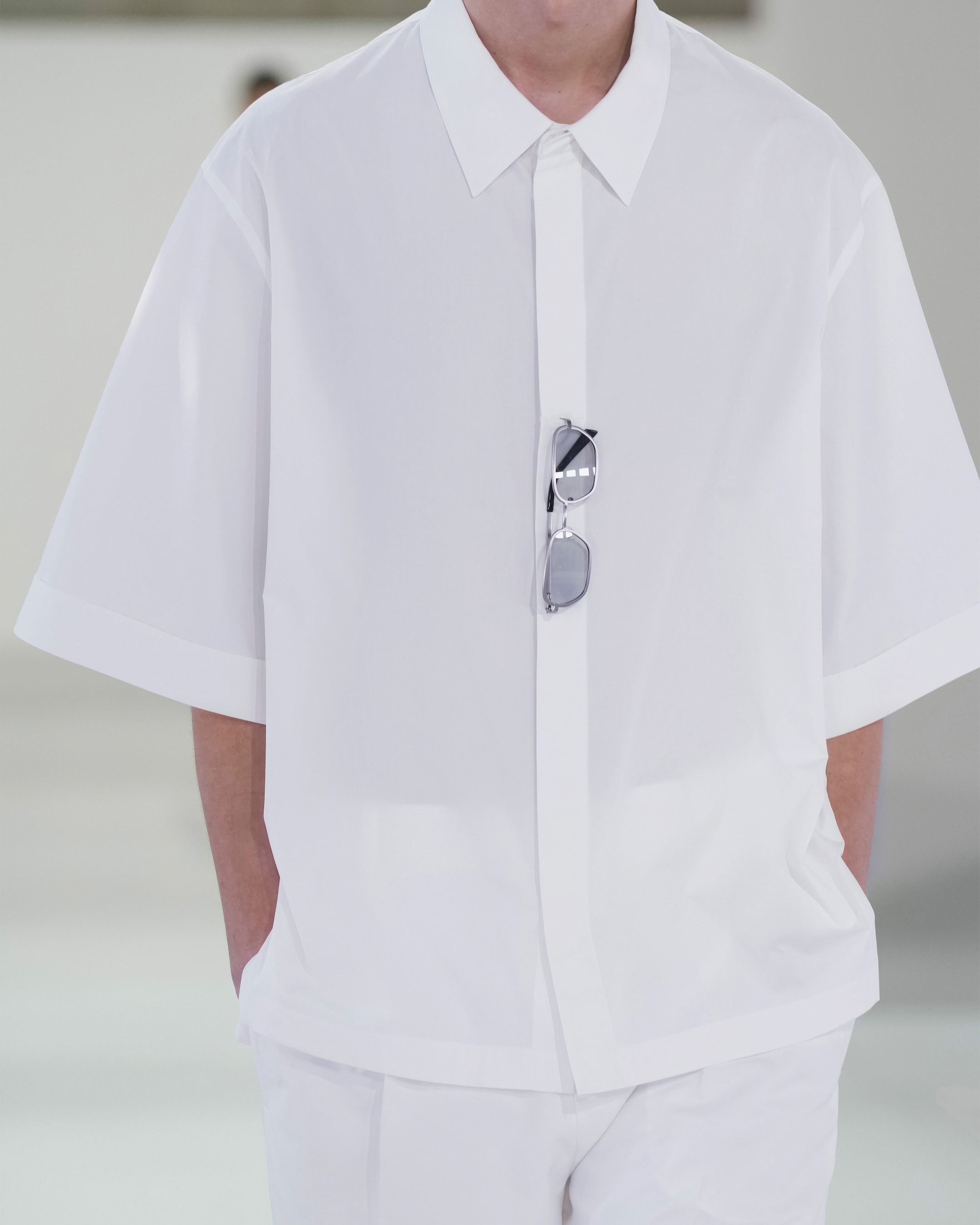 Person wearing a loose white shirt with sunglasses hanging from the collar, their hands in their pockets against a light background.