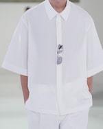 Placeholder for: Person wearing a loose white shirt with sunglasses hanging from the collar, their hands in their pockets against a light background.