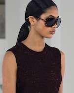 Placeholder for: Person with long dark ponytail wearing large sunglasses and a sleeveless, dark textured top, standing indoors with a neutral expression.