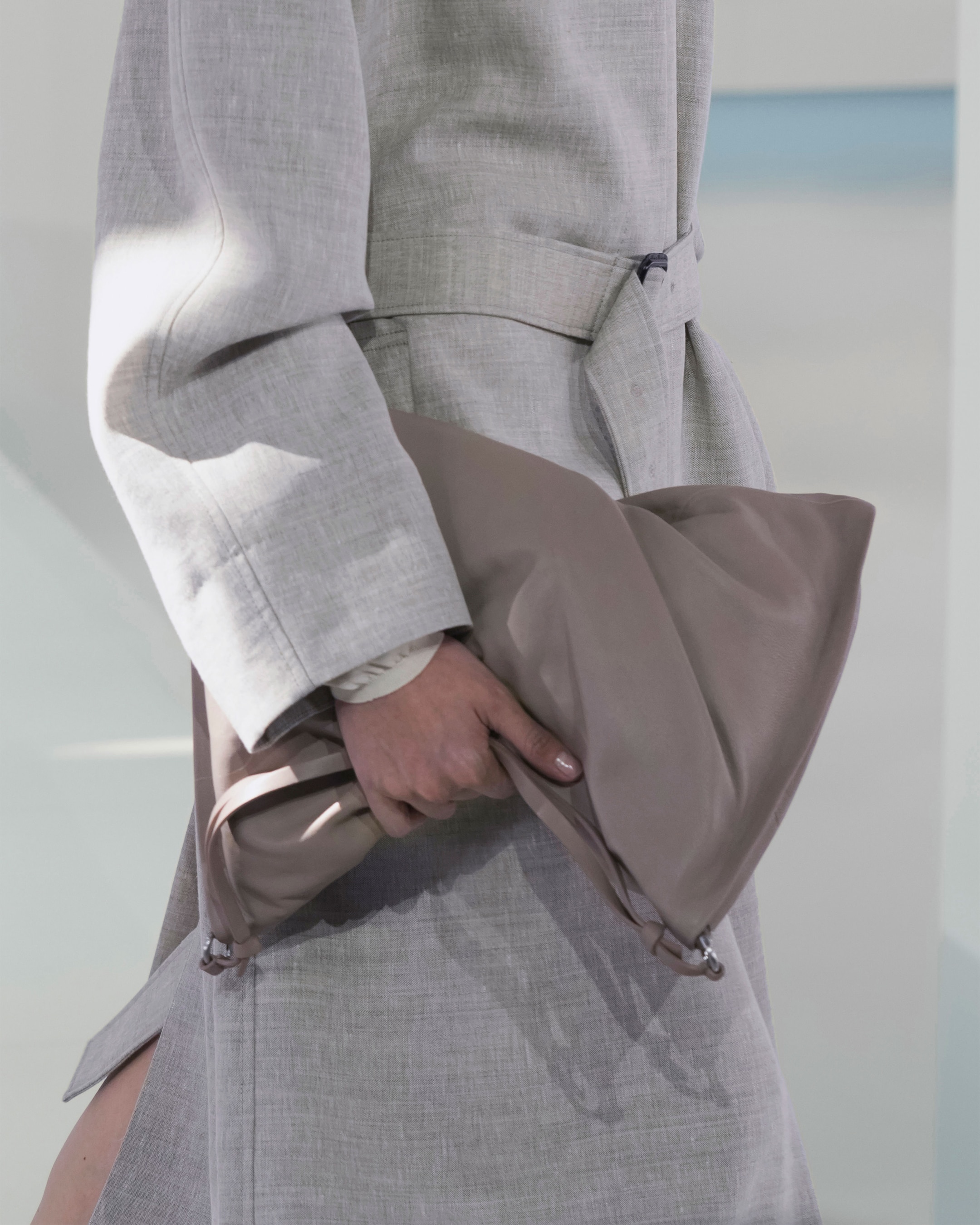 Person in a light gray coat holds a large, soft taupe clutch handbag, with a blurred light background.